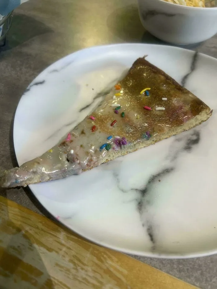 Slice of dessert pizza with icing and rainbow sprinkles on a white marble plate, an example of dishes that are just stupid.