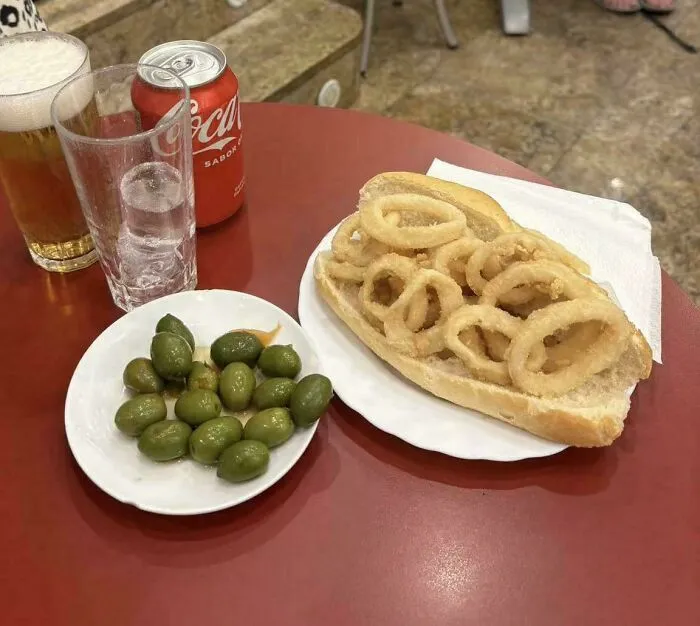 Fried calamari rings served in a sandwich with olives, a glass of beer, soda can, and water on a red table, unusual dish.
