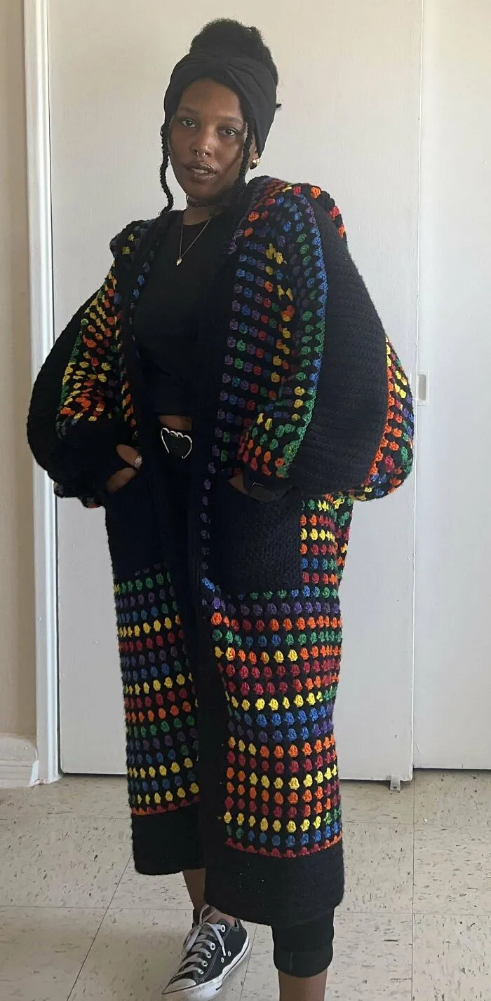 Person wearing a colorful crochet cardigan with vibrant rainbow patterns standing indoors against a plain wall.