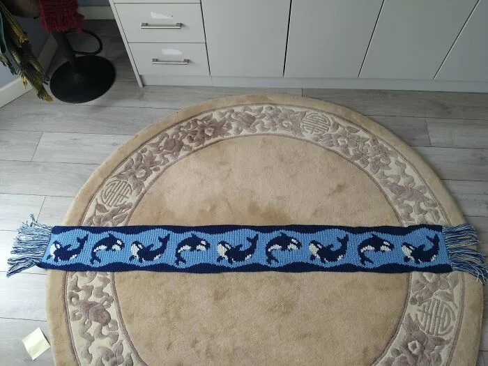 Crochet scarf with repeating orca whale pattern laid out on a round beige and floral rug indoors.