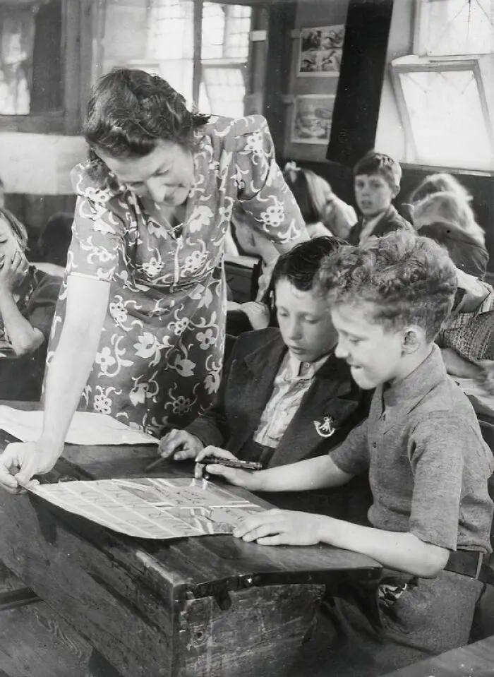 Teacher assisting two boys at a wooden desk in a classroom, a historic scene of schools worldwide during World War II.