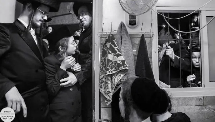 Black and white street photo showing a group of people interacting in a confined space with bars on the window.