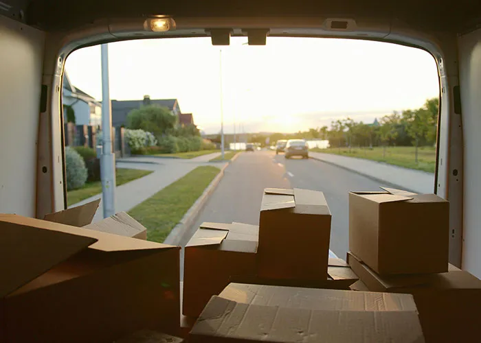 View from inside a moving van filled with packed boxes driving through a suburban neighborhood at sunset.