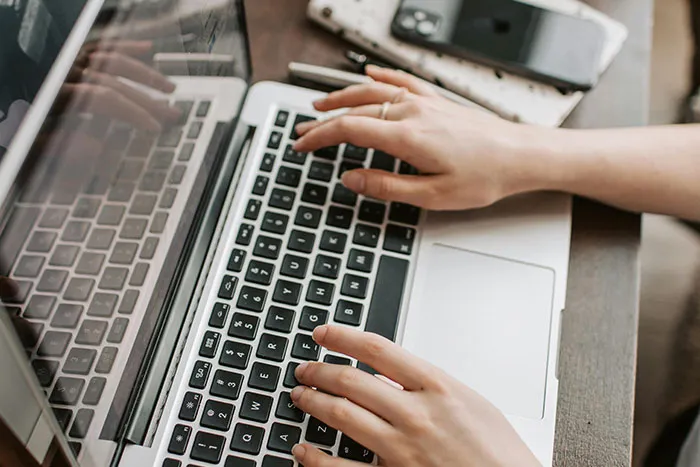 Hands typing on laptop keyboard with phone and keys on wooden desk, illustrating microfeminisms discussion online.