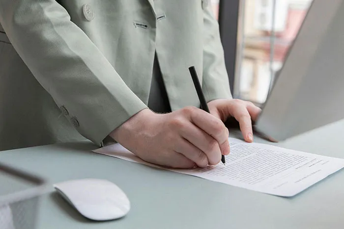 Person in a green suit jacket writing on a document with a pen, illustrating microfeminisms and social commentary.