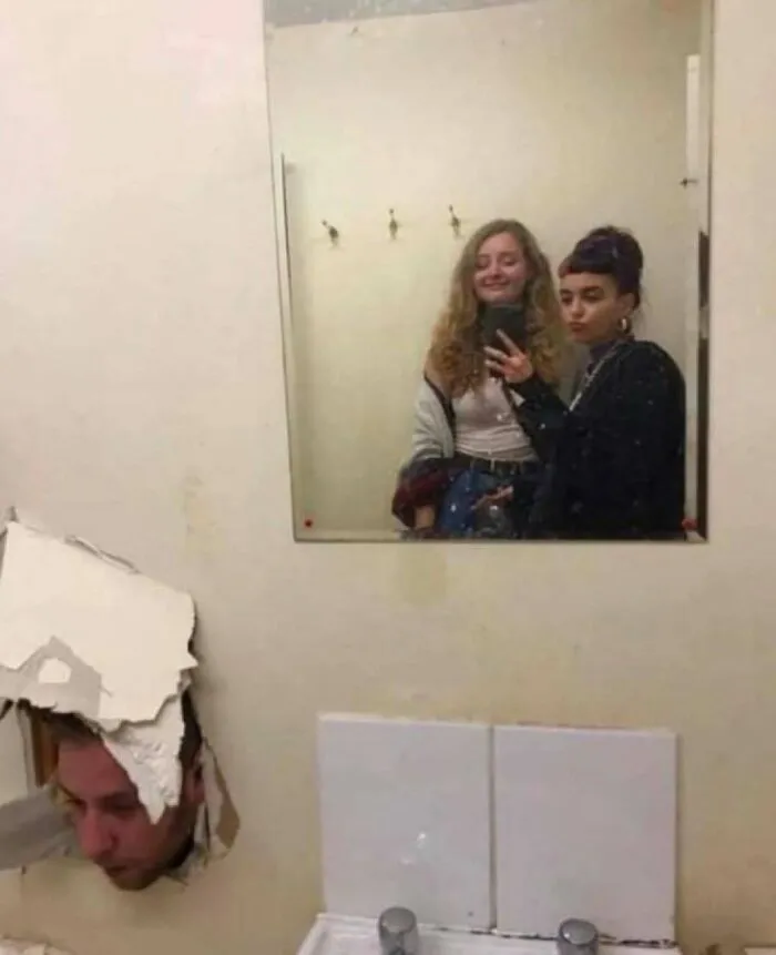 Two women taking a mirror selfie while a man’s head breaks through the wall in a confusing and strange pic.