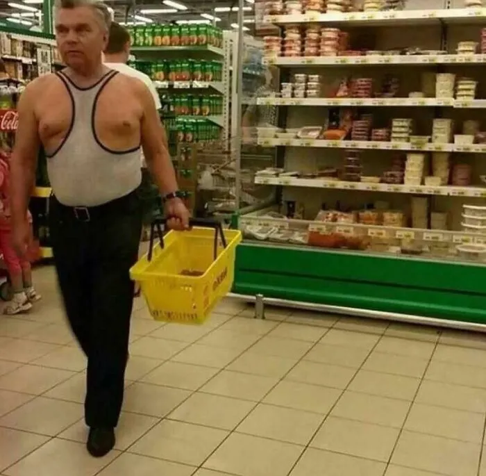 Man wearing an unusual sleeveless shirt walking in a grocery store aisle, holding a yellow shopping basket.