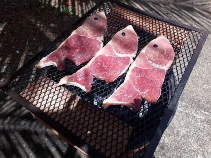 Three pieces of raw meat shaped like fish with olive slices as eyes placed on a metal grill outdoors, bizarre visual.