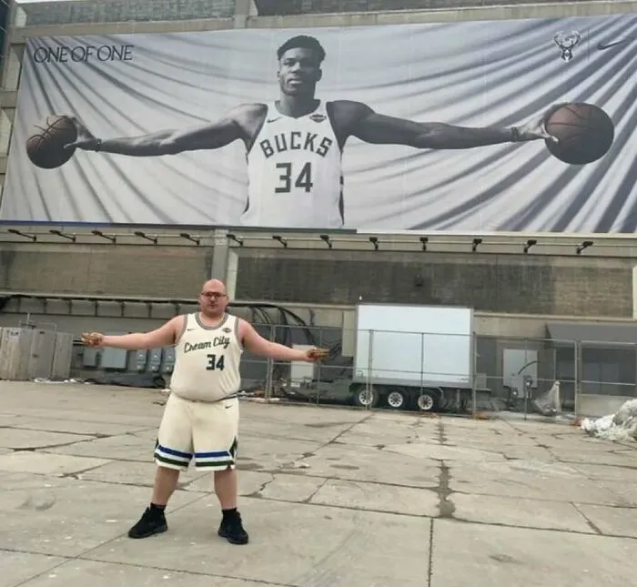 Man in basketball jersey mimics large billboard of player with arms outstretched holding two basketballs, nerdy and weird memes.