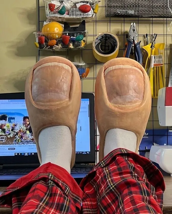 Oversized slippers designed to look like realistic human toes worn with red plaid pants in a workspace setting.