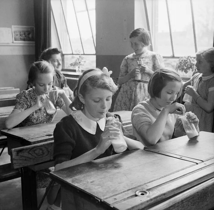 Schoolchildren in a classroom during World War II drinking milk from glass bottles at wooden desks in a historic school setting