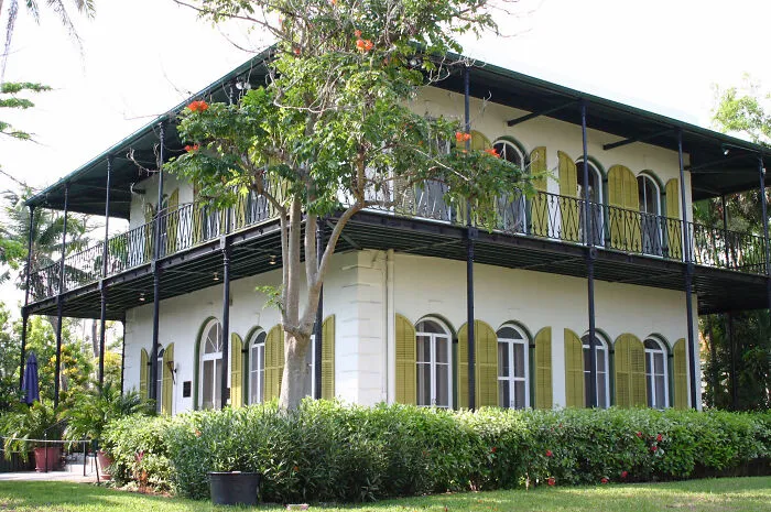 Two-story historic home with wraparound balcony, arched windows, and green shutters surrounded by lush greenery.