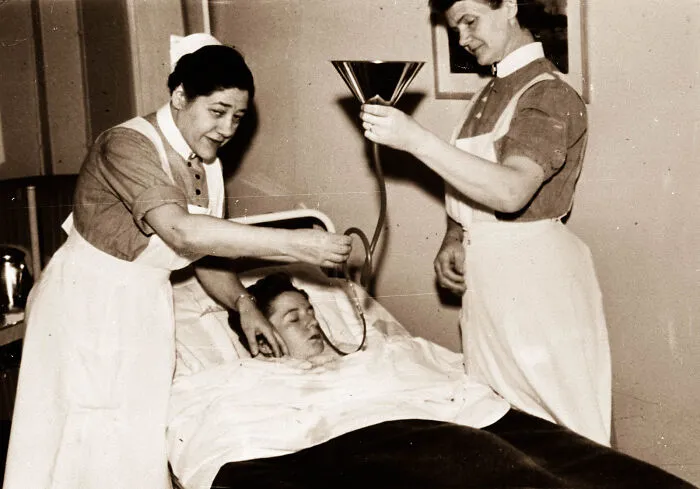 Two nurses using an unsettling medical device on a patient in bed, highlighting vintage medical equipment from the past.