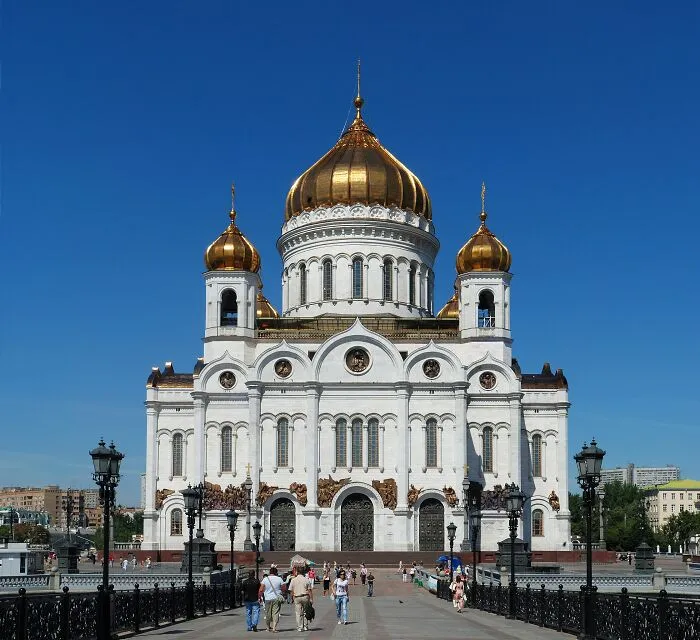 Historic building with gold domes and white facade, surrounded by people and street lamps under a clear blue sky.