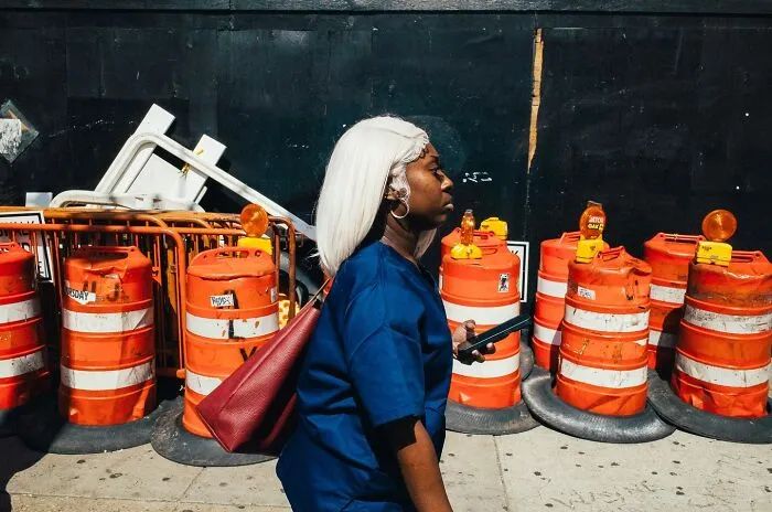 Woman with white hair walking past orange construction barrels in NYC street, showcasing powerful street moments photography.