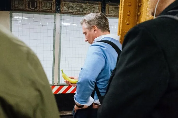 Man in a blue sweater holding a banana on a NYC subway platform, captured in a powerful street moment photograph