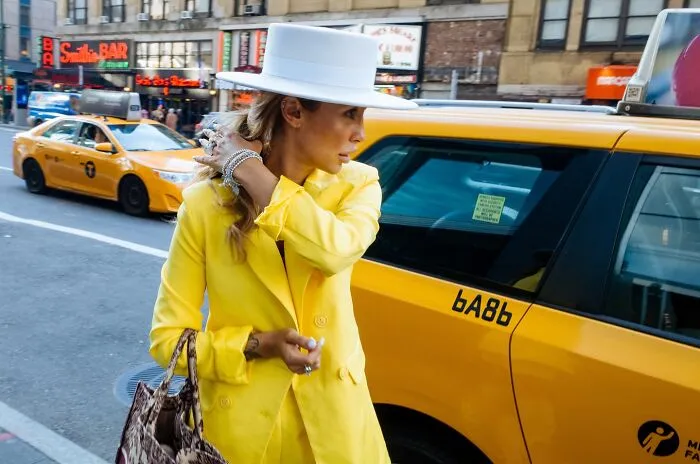 Woman in yellow coat and white hat standing next to a yellow taxi cab in NYC, capturing powerful street moments.