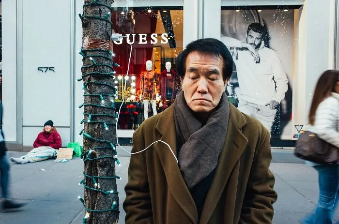Man in brown coat standing on busy New York City street with holiday lights on tree and pedestrians passing by.