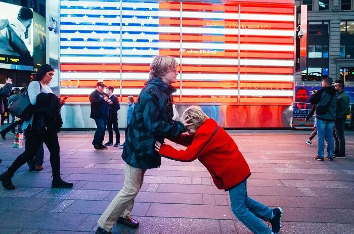 Two children playing energetically in front of a large illuminated American flag in a busy NYC street at night.
