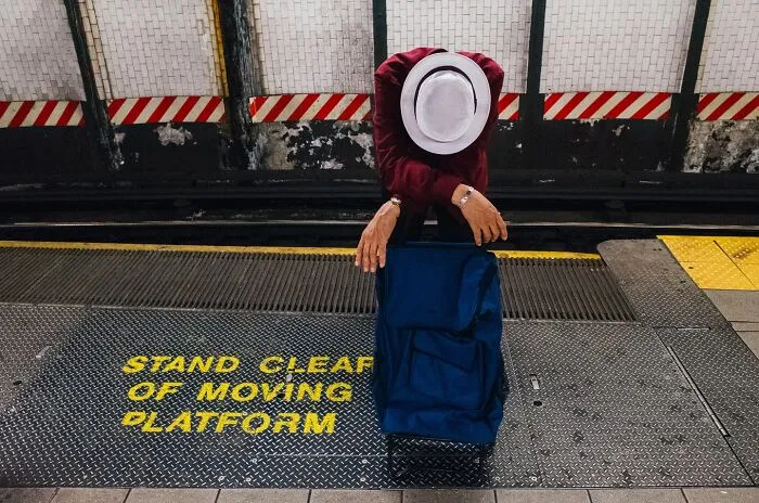 Person in white hat leaning on blue bag on NYC subway platform near a stand clear of moving platform warning.