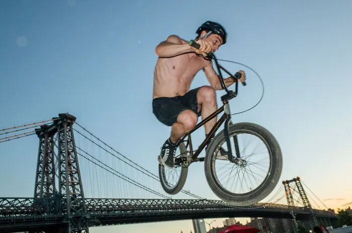 Shirtless cyclist performing a BMX stunt mid-air near an iconic NYC bridge, captured in a powerful street moment.