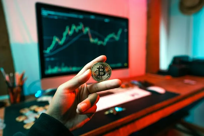Hand holding a bitcoin coin with a stock market chart on a computer screen, reflecting themes of what makes a man unattractive.