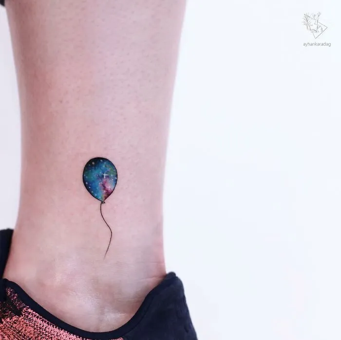 Small ankle tattoo of a colorful galaxy balloon with a thin black string on light skin, one of the best tattoos trending now.
