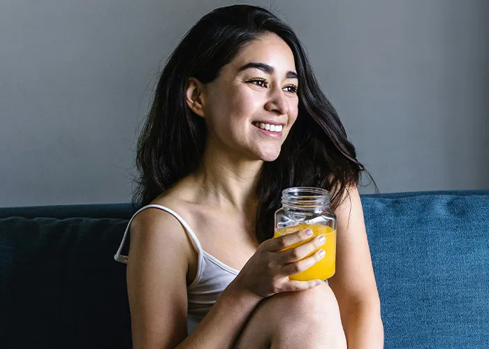 Young woman smiling and sitting on a couch holding a jar of orange juice, doctors wish more people knew health facts