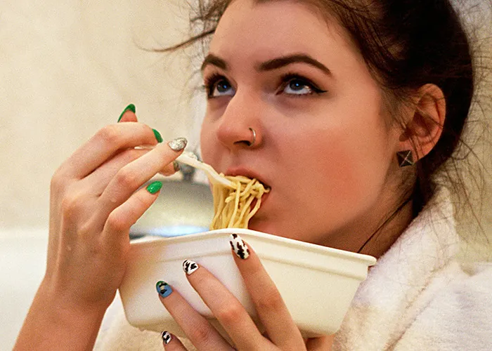 Young woman eating noodles from a container, showing the risks associated with processed foods as death machines warning.