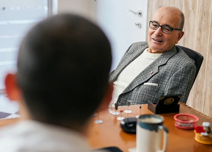 An elderly man wearing glasses and a checkered jacket talking to a doctor in an office about health concerns.