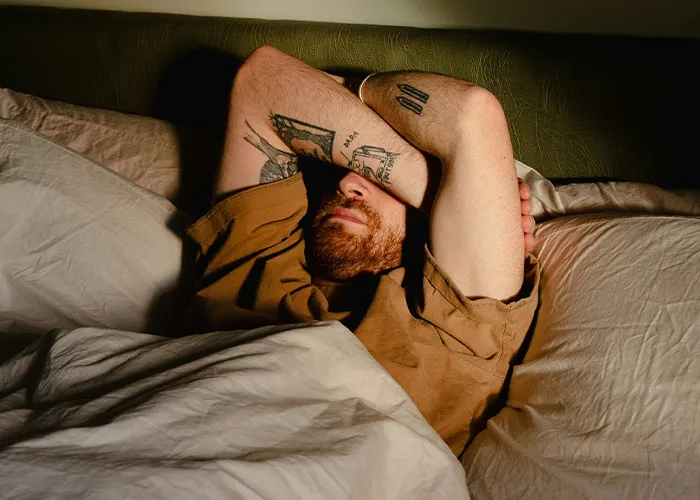 Man with tattoos covering face with arms, lying in bed looking exhausted, illustrating doctors' warnings on health risks.