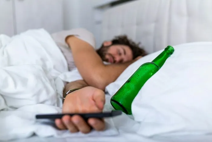 Man lying in bed with beer bottle and phone, illustrating traits that make a man instantly unattractive to women.