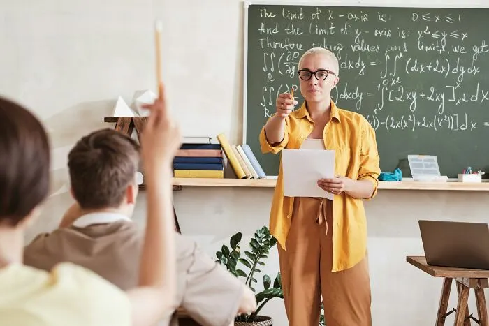 Teacher in yellow shirt teaching math formulas on chalkboard while students raise hands in classroom setting focusing on arrogance.