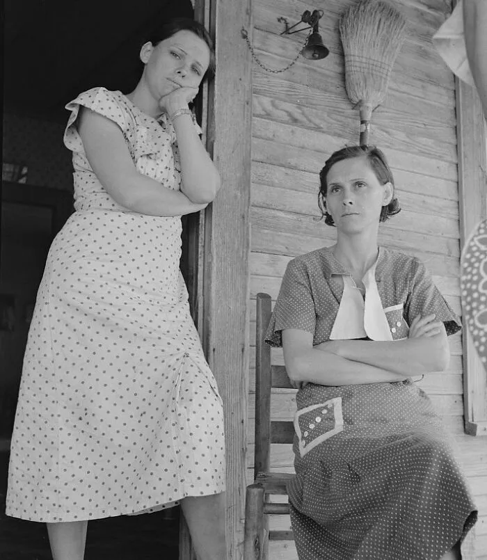 Two women in polka dot dresses on a wooden porch during the Great Depression, showing worry and fatigue.