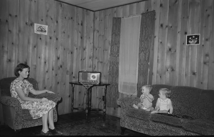Woman in a floral dress sitting in a living room with two children on the couch listening to a radio during the Great Depression era.