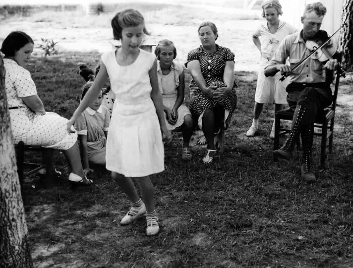 Family in a Great Depression photo with a girl dancing, women watching, and a man playing the violin outdoors.