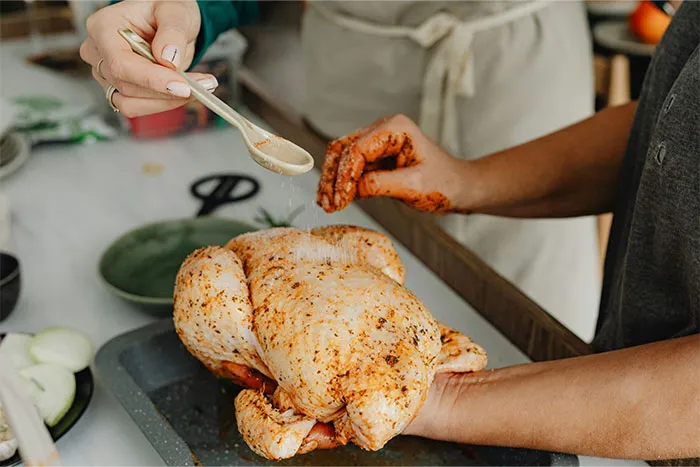 Two people seasoning a whole raw chicken with spices and cooking secrets in a kitchen setting.