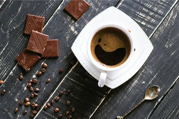 Cup of coffee on a saucer with pieces of chocolate and scattered coffee beans, illustrating cooking secrets and ingredients.