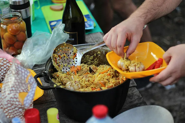 Person serving seasoned rice with secret cooking ingredients from a large pot in an outdoor cooking setting