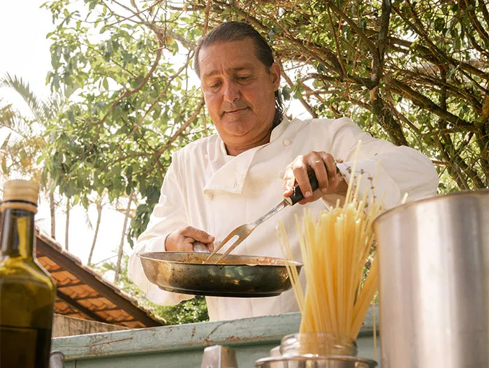 Chef cooking outdoors, sharing cooking secrets and unique ingredients while preparing a dish with spaghetti nearby.