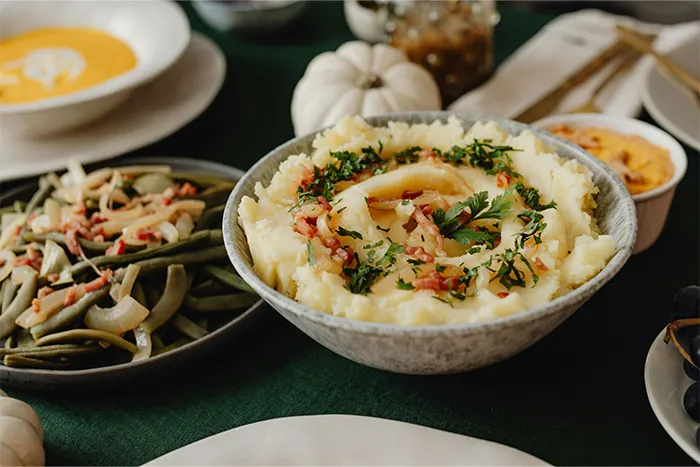 Bowl of mashed potatoes garnished with herbs and bacon, with plates of green beans and soup nearby, sharing cooking secrets.