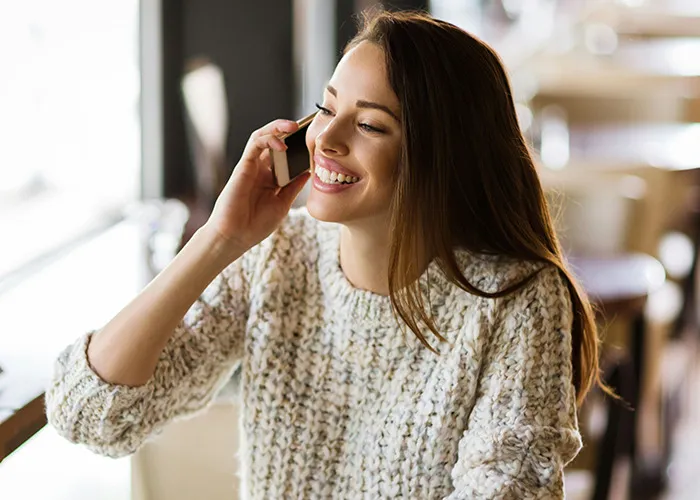 Young woman in a cozy sweater smiling while talking on the phone, illustrating savage insults conversation concept.