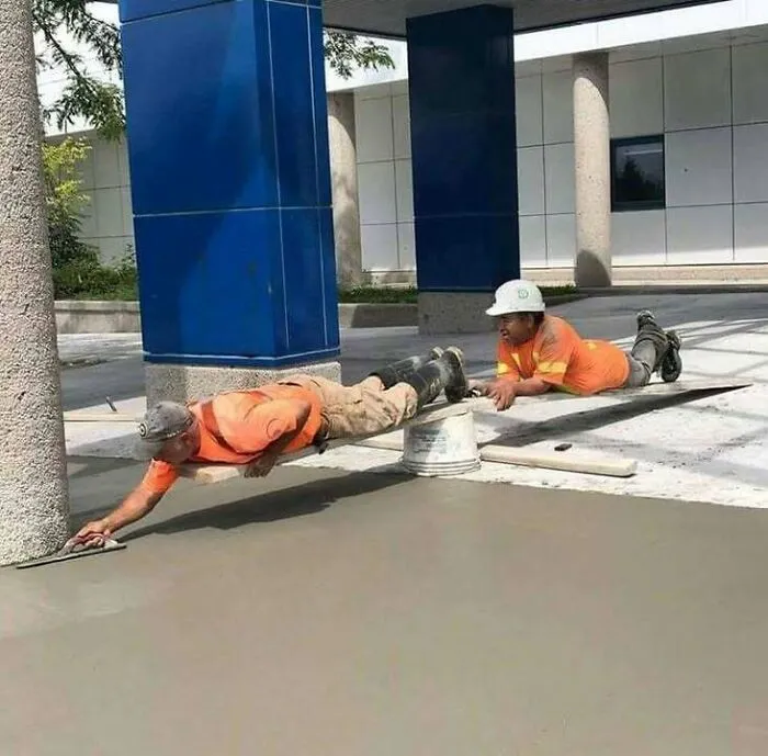 Two construction workers in orange shirts making hilariously dumb mistakes while smoothing concrete at a building site.