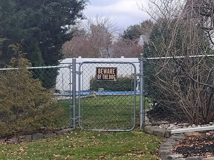 Chain link gate with beware of the dog sign, surrounded by trees and bushes in a neighborhood setting.
