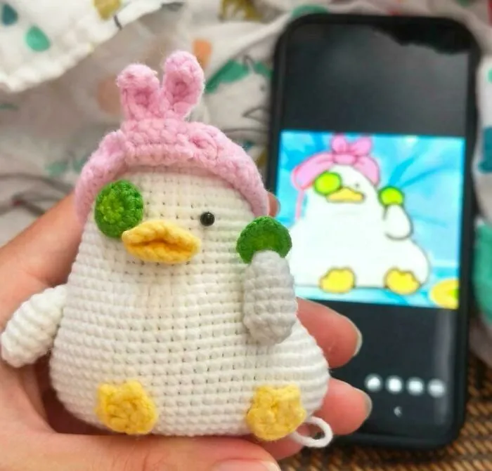 Hand holding a crochet duck with pink bunny ears and cucumber slices, matching an image on a smartphone screen.