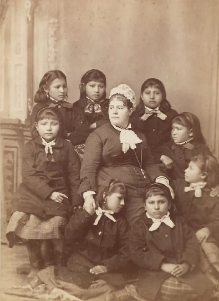Native American boarding school children from the 1910s, dressed in uniform with a teacher in a formal studio portrait.