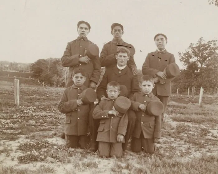 Group portrait of Native American boys and a teacher dressed in early 1900s uniforms at a boarding school.