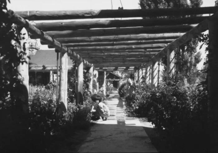 Black and white photo of Native American children under wooden beams at a boarding school garden walkway in the 1910s.