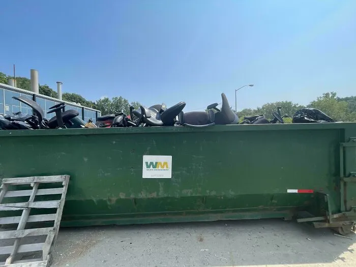 Large green dumpster filled with discarded chair parts representing harsh realities of consumerism waste.