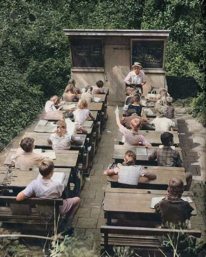 Outdoor classroom with children sitting at wooden desks, a teacher instructing, showcasing interesting historical school setup.